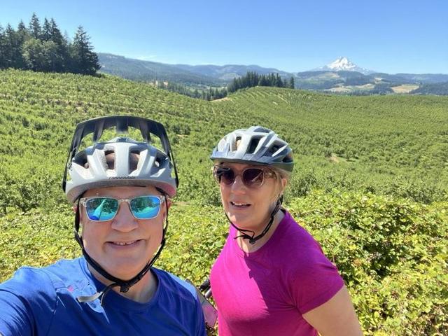 Mark and Lisa Marie Boyd in bike helmets, taking a selfie in front of rolling orchards with a snow-capped mountain behind