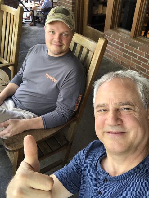 Selfie of Mark Boyd giving a thumbs up on a porch, with his son Brandon seated behind him in a HyreCar shirt and camo cap