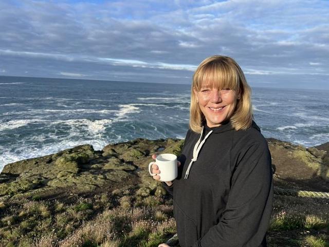 Lisa Marie Boyd holding a coffee mug on a rocky ocean bluff with waves behind her