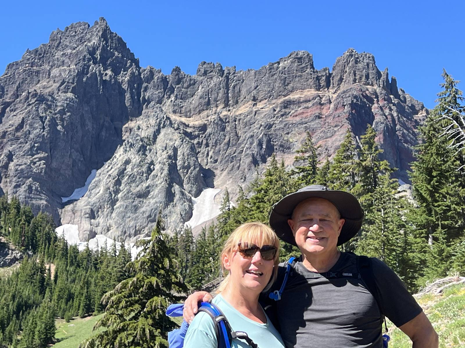 Mark and Lisa Marie Boyd hiking in front of a jagged mountain peak with snow patches and pine trees