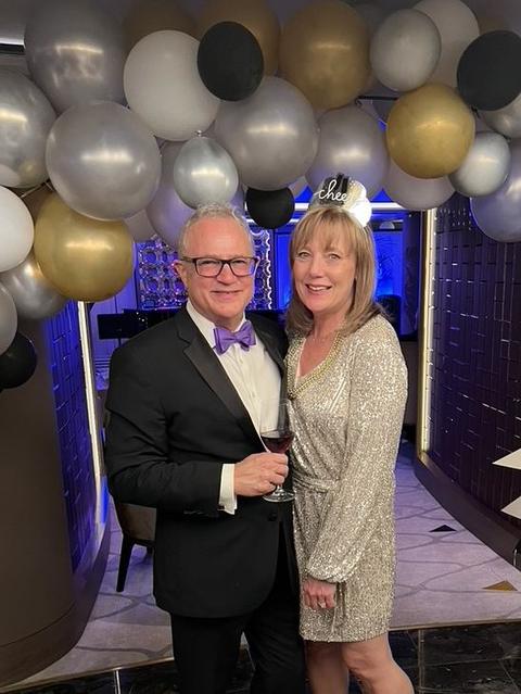Mark Boyd in a tuxedo with a purple bow tie and Lisa Marie in a sequin dress under a New Year's balloon arch