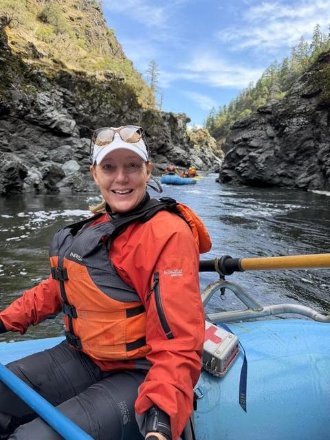 Lisa Marie Boyd rowing a blue whitewater raft through a rocky river canyon, wearing an orange life vest